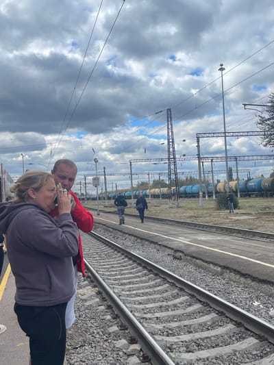 The image portrays a railway environment in Gryazi, Russia, under a sky with prominent gray clouds and patches of blue. In the immediate foreground, on the left, a woman with blonde hair, styled back, is seen wearing a dark gray hooded sweatshirt and dark pants. She holds a bottle with a green cap to her mouth, appearing to drink from it. Her fingernails are painted light green. Standing slightly behind and to her right is a man with a bald or closely shaven head, dressed in a red long-sleeved shirt and white shorts. Both are positioned on a train platform with a yellow safety line visible on the far left. Adjacent to the platform are railway tracks featuring concrete sleepers and gravel ballast.
In the midground, to the right of the foreground tracks, is another platform or walking path. Two male figures are observed walking away from the camera along this path; one wears a dark jacket over light pants, and the other is in dark attire. Further in the background and to the right, a third male figure stands near multiple sets of railway tracks. A long line of stationary freight cars, consisting of blue and yellow tanker wagons, extends across the background. The railway infrastructure includes numerous overhead catenary lines, tall support poles, and lattice towers. A prominent tall light pole with a device mounted on top is visible towards the right midground. Trees and sparse vegetation are discernible beyond the tracks in the distance.