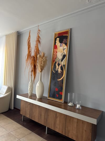 The image shows a close-up of a modern living room in Duszniki, Poland, focusing on a long, low wooden sideboard against a grey wall. On the sideboard, there are two tall, off-white vases. The vase on the left is geometrically textured, while the one on the right is smooth and oval. Dried ornamental grasses, one tall and feathery brown, and another shorter and lighter in color, are arranged in these vases. To the right of the vases, a tall, narrow framed artwork leans against the wall. The artwork depicts a stylized female figure in a vibrant, almost mosaic-like style, with gold, red, black, and white colors dominating. Further to the right on the sideboard are two clear glass cylinders containing white decorative objects, possibly shells or candles. A white armchair is partially visible on the left side of the image, and cream-colored vertical blinds hang in the background, allowing soft light to enter the room. The floor is tiled with large square tiles, a mix of beige and dark brown. The overall impression is one of contemporary interior design, with a focus on natural elements and decorative art.