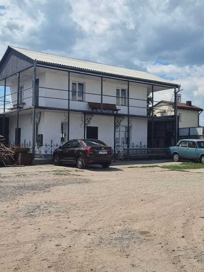 In Pidhaitsi, Ukraine, on a cloudy day, a two-story white building with a corrugated roof stands prominently. A black metal railing encircles the upper story, accessed by an external staircase. Two cars are parked in front of the building on a gravel lot. A dark brown sedan is closer to the viewer, while a light blue older model sedan is parked further away. Piles of wood and other debris are visible to the left of the building. A street sign with Cyrillic lettering, "ВУЛИЦЯ ДАРКОВА 1А" (Dneukova Street 1A), is affixed to the side of the building. The overall scene suggests a commercial or residential property in an outdoor setting.