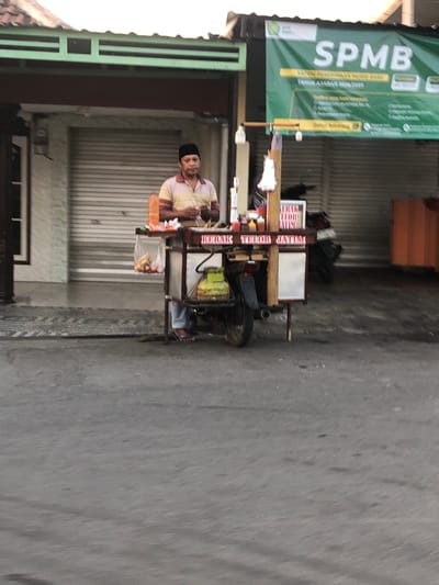A street vendor stands behind his cart, which is attached to a motorcycle, in front of a closed storefront. The vendor, a man wearing a black cap and a striped shirt, is holding something in his hands. The cart has various items on it, including bottles of sauce, raw eggs in a plastic bag, and skewers of food. A gas cylinder is visible on the side of the cart. In the background, a large green banner with the text "SPMB" is displayed, along with other writing in Indonesian. The storefront behind the vendor has a rolled-down metal door. The scene appears to be on a street in Glagah Arum, Indonesia.