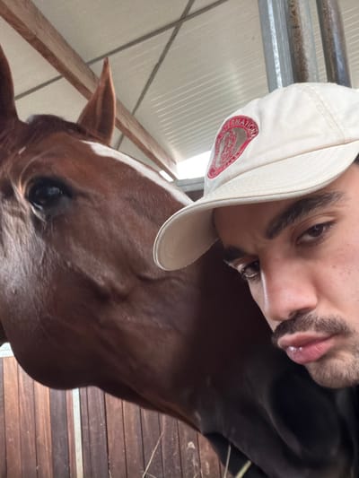 The image displays a close-up, selfie-style shot of a male individual and a horse within what appears to be a stable or barn in Enghien, Belgium. The left side of the frame features the head and upper neck of a brown horse, exhibiting a dark eye, a pointed left ear, and a distinct white blaze on its forehead. On the right, a male individual with dark hair, dark eyebrows, and a mustache-beard is visible, looking directly towards the camera with his lips slightly parted. He wears a light-colored baseball cap featuring a circular red emblem with text, possibly "INTERNATIONAL." The background indicates an indoor structure, with a white corrugated or panelled ceiling and visible wooden beams and metal supports above, transitioning to vertical wooden planking suggestive of a stall partition lower down. Minimal hay or straw is present at the bottom of the frame.