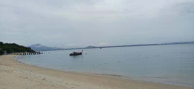 The image depicts a serene coastal scene in Ao Wiang Wan, Thailand. A sandy beach occupies the foreground, meeting a calm, light blue body of water. In the middle ground, a long, narrow boat with a dark hull and visible wooden structure is anchored. Further out, a series of buoys are strung together, marking a boundary in the water. The background features a distant shoreline with lush green trees and gently sloping hills, some of which are shrouded in mist. The sky is overcast with a light grey hue, and a single bird is visible in flight. A pier with multiple wooden pilings extends from the left side of the beach into the water.