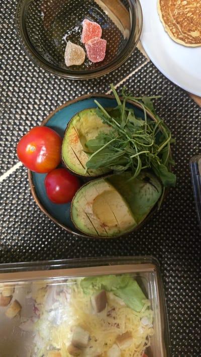 In Ukhta, Russia, on a patterned placemat, a meal is presented. In the upper portion, a clear glass bowl contains three gummy candies—one yellow, and two pink—along with a sprinkle of white salt. Adjacent to this, a white plate holds a single, golden-brown pancake. A teal-colored bowl holds two red tomatoes and a halved avocado, the flesh of which is yellow-green. A small pile of green arugula rests within the avocado halves. In the lower section, a clear plastic container holds a salad composed of shredded yellow cheese, croutons, and leafy green lettuce. The lighting suggests it is daytime, and the setting appears to be a domestic environment.