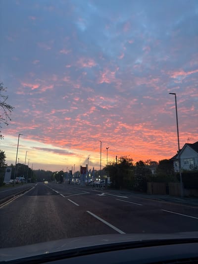 The image depicts a sunset over a road with streetlights in Chester, United Kingdom. The sky is filled with pink and orange clouds. A line of trees and buildings runs along the side of the road, and there are streetlights evenly spaced. The road has white lines marking lanes. The image is taken from a vehicle. The photo is rotated counterclockwise, but the essential elements, such as the city name and the sunset, are easy to identify.