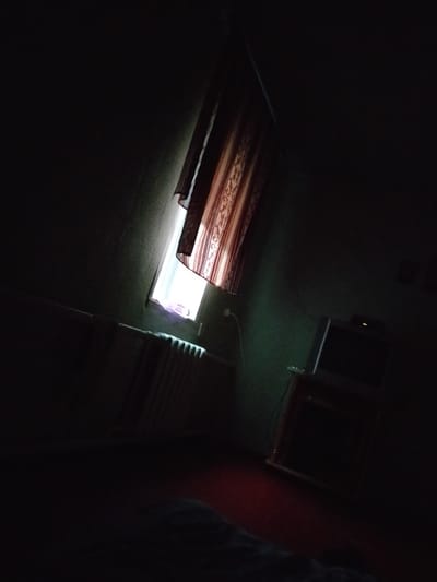 The image shows an interior scene, likely a bedroom or living room, with dim lighting and shadows. A window with a patterned curtain is visible on the left, letting in some light. Below the window, a radiator is present. On the right side of the image, a television set is placed on a wooden stand. The scene is very dark, with only select objects illuminated by the light from the window and the television. The location is identified as Verkhniy Mamon, Russia.