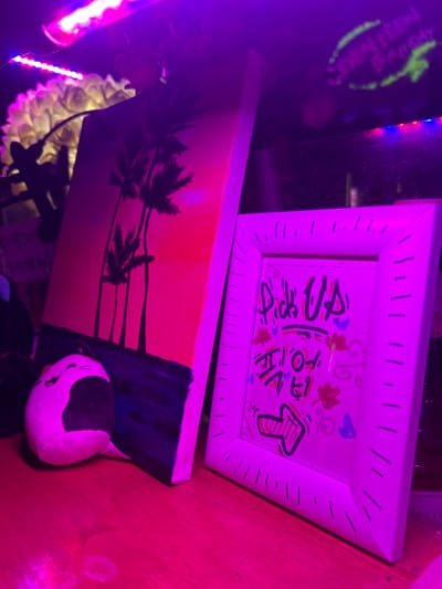This image captures a static scene inside a commercial establishment in Mapo-gu, South Korea, illuminated by prominent purple and pink neon lighting. A reddish-pink strip light is visible at the top of the frame.
On a reflective surface, likely a wooden counter or shelf, three main items are displayed. To the left, a vertical rectangular canvas painting depicts a stylized tropical sunset or sunrise, featuring an orange-red gradient sky above a dark blue foreground. Silhouettes of multiple palm trees stand prominently against the vibrant sky. At the base of this painting, a small, white plush toy with black facial markings and ears, resembling a cat or a stylized food item, lies horizontally on the surface.
To the right of the painting, a white-framed sign stands upright. The frame's inner border is adorned with black dashed lines. The sign's content, handwritten, includes "Pick Up" in English, followed by "픽업" in Korean, and a prominent red arrow outlined in black pointing towards the lower right. Scattered around the text are various small, colorful doodles such as hearts, leaves, and swirls.
In the blurred background, a textured white or light-colored decorative object, possibly a faux plant or coral, is partially visible above the painting. Further to the right, faint green text, possibly "SEVENTEEN Birthday," is visible on a dark surface, suggesting the location may be a themed cafe or event space. No individuals are present in the scene.