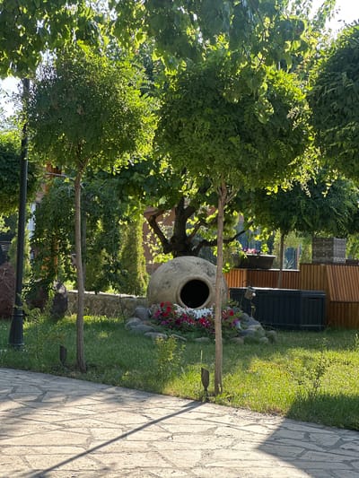 In Ashtarak, Armenia, this image captures a sun-drenched garden area. In the foreground, a stone pathway is visible, leading into a lush green lawn. Dominating the center of the frame is a large, traditional clay amphora, partially embedded in the ground and surrounded by a cluster of colorful flowers. Behind the amphora, a dark, rectangular container is situated, and further back, a wooden fence and seating area are discernible. The garden is shaded by a canopy of green trees, their leaves filtering the sunlight, creating dappled patterns on the ground. The scene appears tranquil and well-maintained, suggesting a private garden or a quiet corner within a larger establishment.