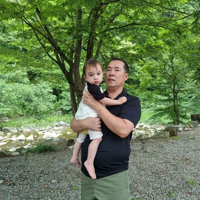 An adult male wearing a black short-sleeved collared shirt and green cargo shorts holds a young child in his arms. The child is wearing a white and black short-sleeved shirt and striped shorts. They are standing outdoors with a body of water and rocks in the background, surrounded by lush green trees. The scene appears to be in a natural park or wooded area. The man is looking towards the camera, while the child is looking slightly off to the side.