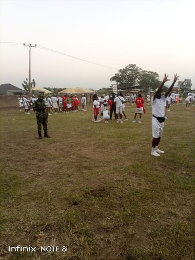 A wide shot captures a field setting with a group of people. Most are dressed in white shorts and shirts, while others wear red tops, and a person in camouflage stands apart. One individual in the foreground raises both hands in the air. A volleyball net is visible in the background, suggesting a sports activity. The sky is overcast, and utility poles are seen in the distance. A phone brand watermark is present at the bottom.