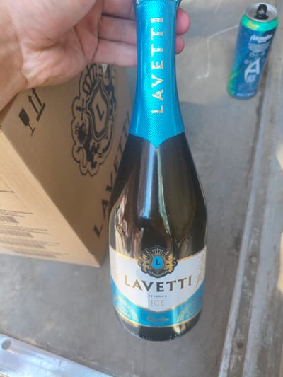 A bottle of Lavetti Ice sparkling wine is held by a person's hand, partially obscuring a cardboard box with a fragile shipping symbol. In the background, a blue aluminum can with the logo "Adrenaline Rush" and "A ICE" is visible. The scene appears to be outdoors, with a concrete or asphalt surface, possibly in a city like Alchevsk, Ukraine. The lighting suggests it is daytime, with shadows cast by the objects.