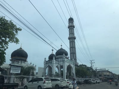 This media file captures a street view in Keudekruenggeukueh, Indonesia, featuring a mosque with striking architecture. The mosque has a prominent tall minaret with intricate patterns and dark domes, characteristic of Islamic religious buildings. In the foreground, several cars and motorcycles are parked or in motion on a paved road, indicating a bustling urban or semi-urban environment. A white minivan with a license plate "BL 1297 NN" is clearly visible.
A sign is posted in front of the mosque with Indonesian text, which appears to be a directive or announcement. The visible text reads: "DILARANG MASUK LOKASI MASJID YANG TIDAK BERBUSANA MUSLIM DAN WANITA BERPAKAIAN KETAT," which translates to "PROHIBITED TO ENTER MOSQUE LOCATION IF NOT DRESSED IN MUSLIM ATTIRE AND WOMEN WEARING TIGHT CLOTHING." This sign provides a cultural and religious context to the location.
The scene is under an overcast sky, suggesting a cloudy or possibly rainy day. The lighting is diffused, and there are no distinct shadows, reinforcing the impression of a somber or muted atmosphere. The presence of power lines crisscrossing the sky is a common urban element. The overall impression is of a typical day in a populated area of Indonesia, with the mosque serving as a central landmark, underscored by the visible signage that guides visitors' behavior.