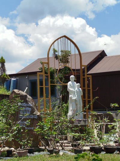 The image depicts a statue of Jesus with two children, set against a backdrop of a building with a brown, corrugated metal roof. A golden archway frames the statue. In the foreground, a large rock is inscribed with Vietnamese text, and various potted plants are arranged on the ground. The scene is set outdoors under a cloudy sky. The rock inscription appears to be a dedication or prayer, possibly related to religious figures. The statue and the surrounding greenery suggest a religious or memorial site.