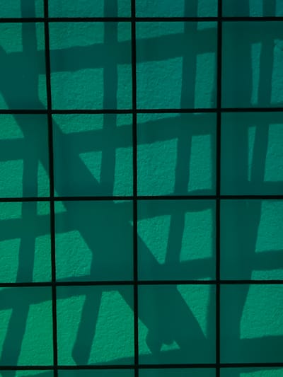 The image displays a teal-colored, textured surface, possibly a wall, located within the city of Derby, United Kingdom. A prominent black grid structure, likely a fence or grate, is positioned in the foreground, casting dark, uniform horizontal and vertical lines across the entire frame. Irregular, angular shadows are cast onto the teal surface, originating from an unseen light source and unidentifiable objects situated between the light and the surface. No individuals, actions, or specific interactions are discernible.