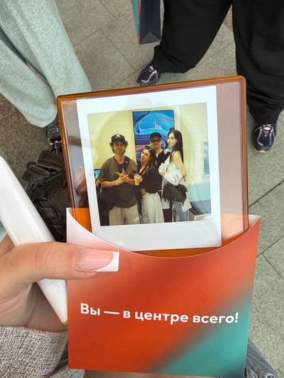 The image shows a close-up of a hand holding a photograph. The photograph features four young people. In the foreground of the photo, a young man wearing a black baseball cap with a white symbol is posing with his fingers in a peace sign. Behind him, a young woman with dark hair is looking at the camera, and another young woman is partially visible. In the background of the photo, a person is seen from the shoulders up. The photograph is held within an orange plastic frame and is partially covered by an orange and teal brochure with text.
The hand holding the photograph has manicured fingernails painted with a French manicure. A white cylindrical object, possibly a vape pen, is also visible in the frame, held by the hand. The background of the image appears to be a tiled floor. The overall scene suggests a casual setting, possibly outdoors or in a public space. The lighting is bright, indicating daytime.
The text on the brochure is in Russian and reads "Вы — в центре всего!" which translates to "You are at the center of everything!".