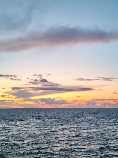 The image depicts a seascape at sunset. The sky is a gradient of colors, transitioning from a pale blue at the top to soft oranges and yellows near the horizon. Several clouds are scattered across the sky, some appearing wispy and others more textured. Below the sky, the ocean stretches to the horizon, its surface marked by gentle ripples. The water is a deep blue, reflecting the subdued light of the setting sun. There are no people or man-made objects visible in the frame, suggesting a natural and tranquil scene. The location is identified as the city of Mologa, Russia.
