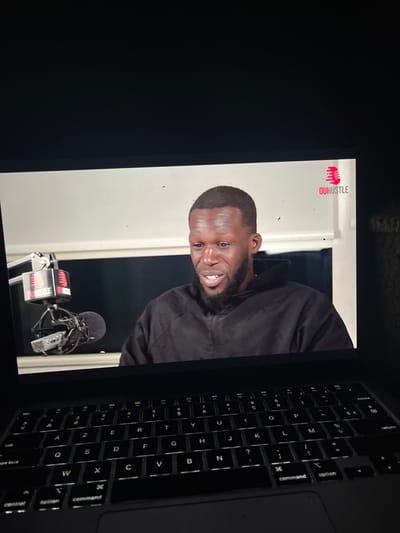 A partially visible laptop screen displays a video of a Black man in a black jacket, speaking into a microphone in a studio setting. The "OUHUSTLE" logo is visible on the left side of the screen. The laptop's keyboard, including keys for "command," "option," and "return," is on the right side of the screen, contrasting with the brighter video image.