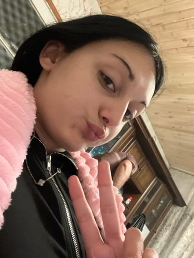 A low-angle selfie depicts a female subject with dark hair and a light complexion, looking directly at the camera. Her lips are pursed in a "kissy face" expression. Her right hand, raised near her face, displays a peace sign (V-sign). She is wearing a black zippered jacket and a light pink, textured, possibly fuzzy, garment draped over her left shoulder and across her chest. A silver-colored necklace with white, four-leaf clover-shaped pendants is visible around her neck. The background shows an indoor environment with light-colored wood paneling on the walls. A dark brown wooden cabinet with multiple doors is partially visible to the right of the subject. An out-of-focus limb, possibly an arm or leg, clothed in a brown fabric, is situated behind the cabinet. A small red and white bottle is visible on a surface beneath the cabinet. The precise location within Lanchkhuti, Georgia, is not discernible from the provided image.