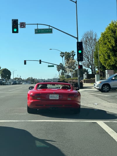 The image shows a red car with a California license plate "9PVY453" stopped at a green light. A street sign reads "Del Amo Circle East". Another street sign says "Madison". Another sign is visible for an Auto Detailing Supplies Store. The sky is clear and blue. Palm trees line the background, and other cars are parked along the street.
