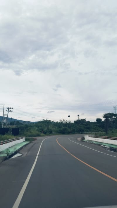 The image presents a daytime scene from the perspective of a vehicle traveling on a two-lane asphalt road in Lonbaroh, Indonesia. The road, marked with a solid white line on the left shoulder and a broken yellow center line, gently curves towards the right, leading into the distance.
On both sides of the curving road, low concrete barriers or railings are visible, painted white with a strip of green at their base. A small puddle of water can be seen next to the barrier on the left.
The surrounding landscape is characterized by abundant green vegetation. Dense trees and bushes line the road and cover rolling hills in the background. On the left side, utility poles with power lines and a visible transformer indicate nearby infrastructure. Further into the mid-ground on the right, two distinctive, tall palm trees with sparse foliage stand above the treeline. A large metal power transmission tower is partially visible in the far right background, partially obscured by foliage.
The sky is overcast with a spread of soft grey clouds, suggesting a cloudy day with diffused lighting, rather than direct sunlight. There are no people, animals, or visible text in the image. The overall impression is one of a peaceful drive through a verdant, semi-rural or natural setting in a tropical climate. The scene captures a moment of travel or observation on what appears to be a quiet road.