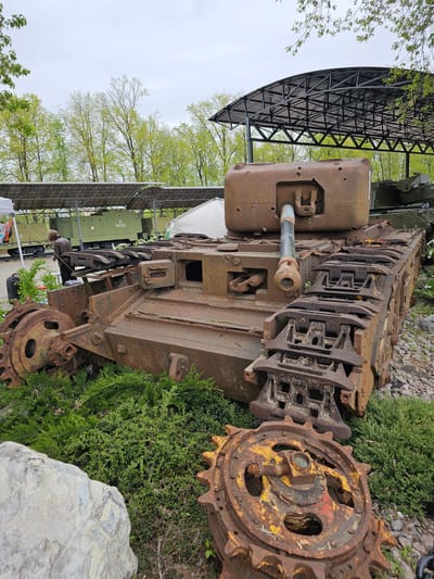 The image captures an old, heavily rusted military tank in the park of Arkhangel’skoye, Russia. The tank is the primary subject, exhibiting significant corrosion throughout its structure, including the turret, gun barrel, tracks, and drive sprocket. The tank sits beside lush green bushes and a large gray rock. In the background, there is a modern, green military vehicle parked under a black metal canopy, and trees with fresh spring foliage reaching towards a cloudy sky.