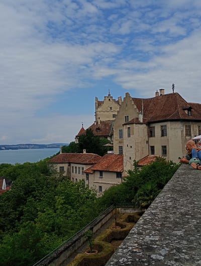 The image depicts a view of Meersburg Castle in Meersburg, Germany, overlooking Lake Constance. The castle, with its distinctive terracotta roofs and light-colored walls, is situated on a hill surrounded by lush green trees. In the foreground, two people are visible from the waist up, leaning on a stone wall and looking at a phone. They are dressed casually. The lake stretches into the distance, with a hazy shoreline visible on the opposite side. The sky is partly cloudy, with patches of blue sky showing through. A well-maintained garden with manicured hedges and a small potted tree is visible on a lower terrace below the castle. The stone wall in the foreground is textured and weathered.