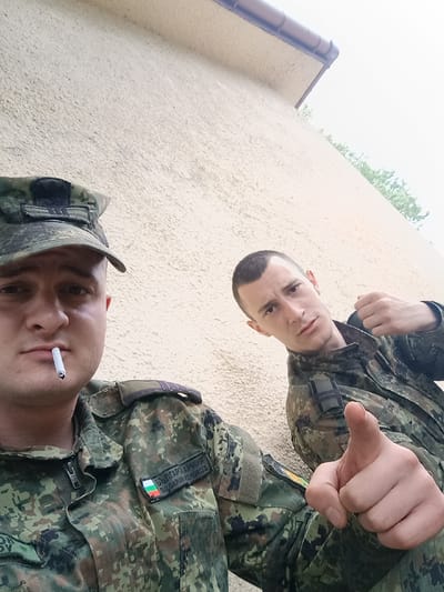 Two men in military camouflage uniforms are in the frame. One man, closer to the camera, is wearing a cap and has a cigarette in his mouth. The patch on his uniform indicates Bulgarian military service. The other man is below him and is also wearing a camouflage uniform. The location appears to be outdoors near a building. It is impossible to confirm location based on the image.