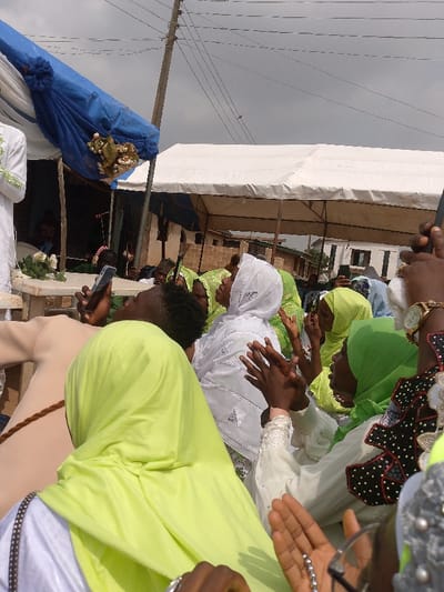 An outdoor gathering is depicted in Ijoko, Nigeria, under an overcast sky. The scene is dominated by multiple individuals, primarily women, positioned in what appears to be a crowd. Many are looking upwards or towards a central, unseen focal point.
In the foreground, a person with dark, short hair and a lime-green head covering (hijab) is visible from the back. To their right, a woman in a white patterned hijab looks upwards, with both hands raised and clasped as if in applause or prayer. Another woman to her right wears a dark green hijab, also with raised hands. Further to the right, a hand adorned with a gold watch and rings is seen holding a smartphone.
On the left side of the frame, a person in white attire stands partially visible behind a white stand or table decorated with white roses. A blue fabric canopy, adorned with artificial flowers, hangs overhead. In the mid-ground, a white tent structure is visible, extending towards the right. Utility poles and overhead power lines traverse the upper portion of the frame against the cloudy sky. In the background, various residential or commercial buildings constructed from light-colored bricks are visible. Several other individuals in head coverings of varying green and white hues are scattered throughout the crowd, some also with hands raised. One person in the mid-ground holds a smartphone upwards, possibly recording the event.