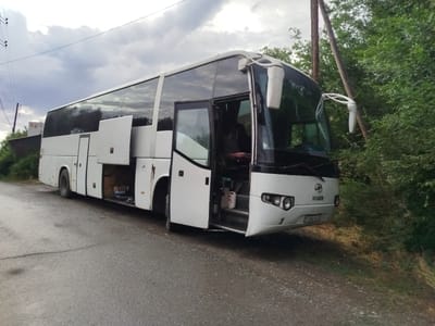A white Higer bus is parked on the side of a paved road in Kabanbay, Kazakhstan. The bus's front passenger door is open, revealing the steps leading into the cabin. Several storage compartments along the side of the bus are also open, exposing their contents. The bus is surrounded by lush green trees and vegetation, with power lines visible in the background. The sky is overcast with dramatic clouds. The license plate of the bus is visible and reads "856 ZX 05".