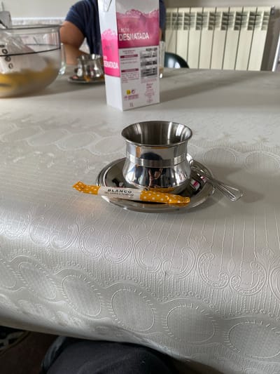 The image captures an indoor scene in Villaviciosa, Spain, featuring a table set for a beverage or meal preparation. In the immediate foreground, a reflective silver-toned metallic cup, distinguished by a textured band, sits on a matching saucer. A silver spoon rests on the saucer next to the cup. A yellow packet with white polka dots, labeled "BLANCO AZÚCAR del campo a tu hogar" (BLANCO SUGAR from the field to your home), is positioned on the saucer. The table is covered by a white, embossed tablecloth exhibiting intricate patterns including swirls, vertical lines, and circular motifs. In the midground, a white 1-liter carton of "Leche DESNATADA" (Skimmed Milk) with a pink graphic is prominently displayed. The background is out of focus, revealing a person's arm clad in a blue shirt. To the left, a clear glass mixing bowl contains a yellow liquid and a partially submerged utensil. Further back, a smaller metallic cup and saucer are visible. On the far right, a white radiator is affixed to a light-colored wall, and a dark chair is partially discernible. The bottom of the frame shows the dark-clothed lap of a person seated at the table.