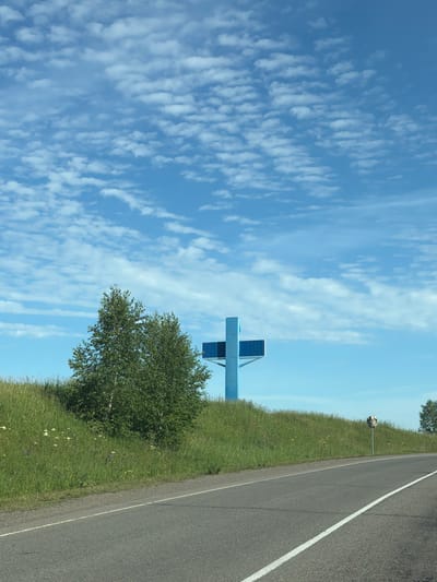 A tall, bright blue cross-shaped structure stands against a sky dotted with wispy clouds. The cross appears to be an advertisement or sign, with blue panels forming its horizontal and vertical bars, and a smaller circular blue element at the intersection. It is positioned on a grassy hill. To the left of the cross, a green, leafy tree grows. The hill slopes down to a paved road in the foreground, which has a white line marking its right edge. The environment is rural, with a clear sky and verdant vegetation. There are no people visible in the image. The location is Ozero Uchum, Russia.