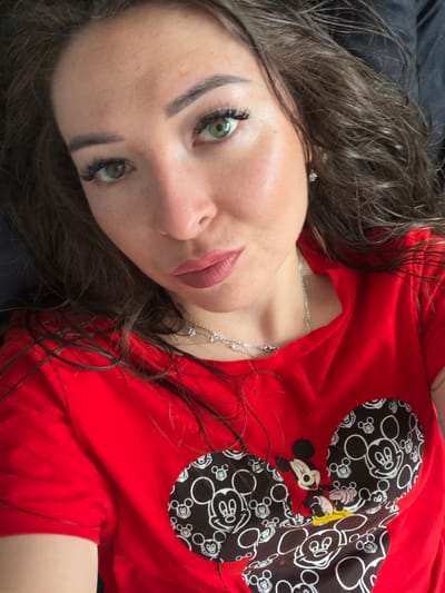 The image is a close-up, high-angle selfie of a woman with long, dark, wavy hair, green eyes, and a fair complexion. She is wearing a red t-shirt with a Mickey Mouse graphic. Her lips are painted with a matte pink lipstick. She is also wearing a thin silver necklace. The background appears to be a dark, possibly black, surface. There is no discernible information in the image that would suggest a location in the city of Япрыково, Russia.