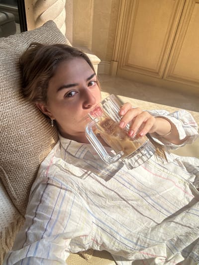 A female subject is depicted in a reclined position, looking directly at the viewer, while drinking from a clear, ridged glass mug containing an iced, milky brown beverage. She wears a light-colored, long-sleeved button-up shirt with thin multicolored stripes. Her left earlobe displays a small drop earring with two linked circles and a pearl-like bead, and a thin chain bracelet is visible on her right wrist. Her fingernails are painted a light shade. She rests against a textured, beige cushion on what appears to be a light-colored couch or lounge chair. The background features a warm-toned interior, including a twisted architectural column on the left and light-colored wooden cabinetry with decorative molding and dark hardware on the right. Strong sunlight illuminates the scene, casting distinct shadows. No visual identifiers are present to ascertain the specific location within the city of Montgomery, United States.