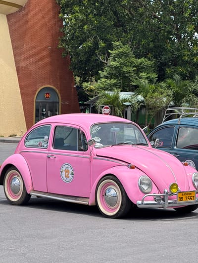 A bright pink Volkswagen Beetle is parked on a paved road, with a portion of a brick building and lush green trees in the background. The car is the main subject, positioned in the foreground and taking up most of the frame. It has chrome accents, white-rimmed tires, and a license plate with yellow background and black text that reads "กศ 7557". There are stickers on the car, including one on the side door that says "Lao Volkswagen Caravan 12" and another on the rear window with "SAVAN VW Club". To the right of the pink car, a dark green Volkswagen Beetle is partially visible, also with a roof rack. In the background, a red brick building with an arched doorway is visible, along with dense foliage and palm trees, suggesting a tropical or subtropical climate. A red STOP sign is also visible above the cars. The lighting suggests it is daytime with bright sunlight. The location is identified as Lao Bao Commune, Vietnam. There are no people visible in the image.