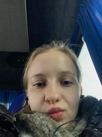 The image presents a close-up view of a young Caucasian female seated within a vehicle in Khodosivka, Ukraine. She is facing directly forward, her gaze meeting the camera, and her lips are pursed. Her hair, light in color, exhibits several prominent flyaways. She has blue eyes. Black wired earphones are inserted into both ears, with their cords visible. She wears a dark, possibly quilted jacket, and a textured, grey, fuzzy or knit garment, possibly a scarf, is wrapped around her neck and lower face. The immediate environment includes a dark blue/grey overhead panel featuring a circular vent or speaker element in the upper left background. To the upper right, a royal blue pleated curtain, partially drawn, covers a window, revealing a bright, diffuse light source beyond. A portion of a blue upholstered seat is visible to the lower left of the frame.