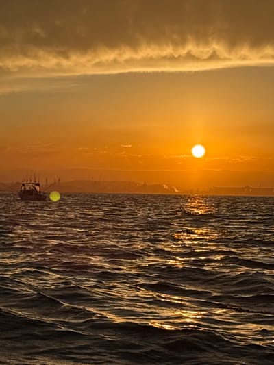 This image presents a marine scene at either sunrise or sunset, observed from the water towards the coastline of Port Kembla, Australia. The sky displays a golden-orange and yellow gradient, with a prominent, bright, circular solar disc located in the upper right quadrant. A distinct, horizontal band of undulating clouds spans the upper portion of the sky. The foreground and midground consist of dark, choppy water, reflecting the sun's light in a shimmering path extending from the horizon towards the observer. A white-hulled fishing vessel, equipped with multiple fishing rods, is positioned in the mid-left. Several silhouetted individuals are visible within the vessel's cabin. The distant horizon reveals the industrial infrastructure of Port Kembla, including cranes, structures, and what appear to be industrial emissions or steam. A large vessel or fixed structure is discernible towards the right horizon. A spherical, patterned yellow-green optical artifact is situated to the left of the fishing vessel.