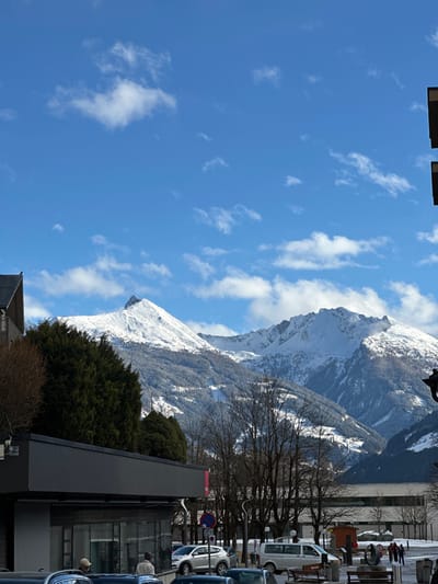 The image depicts a winter street scene in Bad Hofgastein, Austria, with snow-covered mountains dominating the background under a clear blue sky dotted with scattered white clouds.
In the foreground and midground, urban infrastructure is present. On the far left, a portion of a traditional building with a dark pitched roof is visible. Adjacent to it, a modern commercial building with a dark facade, a flat roof, and large glass windows occupies the lower-left section. Two individuals are partially visible inside or directly in front of these windows; one wears a light-colored hat. A dense cluster of dark green coniferous trees stands behind this building, partially obscuring the lower mountain slopes.
A street or parking area runs across the mid-ground, featuring several parked vehicles including a silver SUV, a white sedan, and a white van. Bare deciduous trees with dark branches line this area, interspersed with streetlights. A blue circular traffic sign with a red diagonal line (indicating a parking or stopping restriction) is visible. Further to the right, a pedestrian plaza is observed, covered in light snow, containing outdoor benches, a small brown wooden kiosk-like structure, and a dark, abstract outdoor sculpture. Several people are visible walking or standing within this plaza.
In the mid-right background, a large, light-colored modern building with multiple horizontal window sections is partially obscured by trees and the plaza elements. The entire scene is brightly lit by direct sunlight, evidenced by clear shadows on the snow and buildings. The prominent snow-capped peaks of the Alps rise majestically in the background, with visible forested areas and what appear to be ski tracks or slopes on their lower sections.