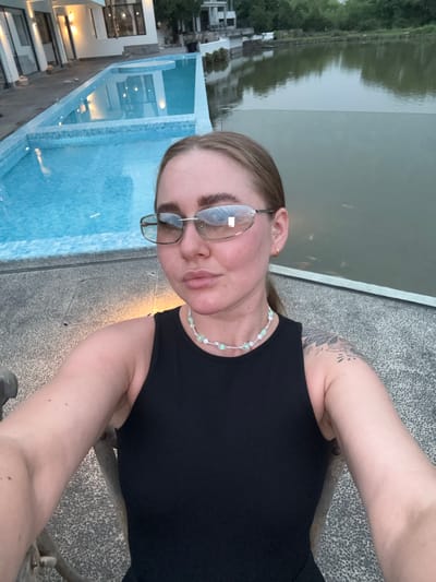 A woman is depicted in an outdoor setting, likely a resort or upscale residence, in the city of Jinhua, China. The woman wears glasses, a black tank top, and a beaded necklace. She has a tattoo on her upper arm. A pool with blue tiles is visible to the left. A body of water, possibly a river or lake, is behind her, with vegetation visible on the opposite bank. Buildings are also present on the far shore. The surface she is positioned on appears to be concrete.