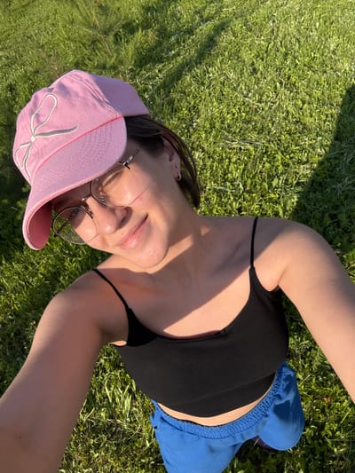 A young woman with dark hair, wearing a pink baseball cap with a white bow detail, glasses, a black tank top, and blue shorts, takes a selfie outdoors. She is smiling and looking at the camera. The background is a sunlit grassy area. There is no indication of the city of Doksha, Russia in this image.