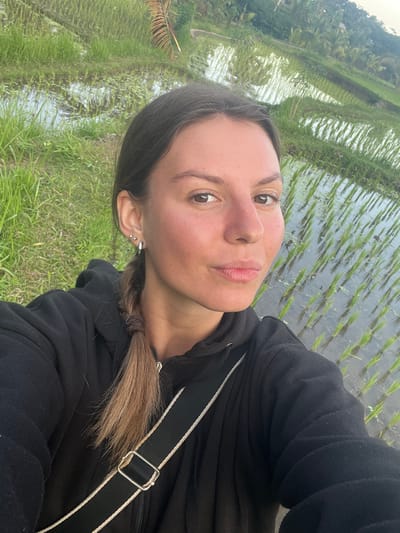 A young woman with brown hair in a braid takes a selfie in front of a lush green rice paddy. The rice paddy, with its neatly planted rows of young rice stalks, is partially flooded with water, reflecting the sky and surrounding vegetation. The woman is wearing a black hooded jacket and a cross-body bag with a black strap and white trim. She has several piercings in her ear. The background shows more of the terraced rice fields, a palm tree, and other tropical foliage, suggesting a rural setting in Tetebatu, Indonesia. The lighting indicates it is either early morning or late afternoon, with a soft, diffused light.