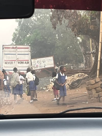 A group of school children wearing uniforms walk along a dusty road. Some are in blue dresses with white tops, while one wears red shorts. Behind them is a parked truck with the words "NEVER WORK ALONE" printed on its side. A sign with illustrations stands nearby, and a pile of rubble is visible in the background. Trees overhang the scene, adding to the atmosphere.