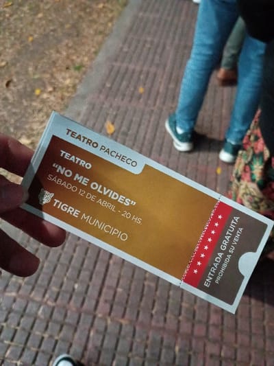 The image shows a hand holding a ticket for a play called "No Me Olvides" at the Teatro Pacheco in Tigre, Argentina. The ticket is gold and white with red text. It says "Entrada Gratuita Prohibida Su Venta" which means "Free entry. Sale prohibited". The location is in the city of General Pacheco, Argentina, with coordinates -34.457 latitude and -58.633 longitude. The event is scheduled for Saturday, April 12th, from 8 pm to 10 pm. The background is a brick walkway, and there are blurred figures of people in the background.