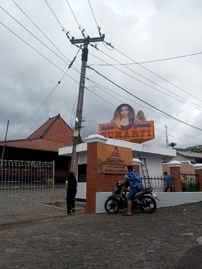 The scene is located in the city of Saing Ta Mawt Lay, Myanmar. An outdoor view under an overcast sky depicts a commercial establishment and street. A prominent orange sign above a white building entrance displays "Ayam Goreng SUHARTI" in blue text, accompanied by a circular portrait of a woman. Below this, an orange banner on a brick wall announces "TELAH DIBUKA TRIAL OPENING RUMAH MAKAN KHAS YOGYAKARTA TANGGAL 23 MARET 2026." To the left of the white building, a traditional-style building with a reddish-brown tiled roof is partially visible behind a grey metal gate. A tall wooden utility pole with multiple power lines stands prominently in the center.
Three individuals are present. On the left, a person in dark clothing walks away from the camera, partially obscured by the partially open grey metal gate. In the mid-ground right, a male wearing a blue and white patterned shirt and blue shorts stands next to a black motorcycle, positioned as if dismounting or preparing to mount. On the far right, a third male, wearing a light blue shirt, is partially visible behind a brown metal fence, near a silver ladder leaning against the white building. The foreground consists of an uneven paved surface. The sky is largely covered by grey clouds.