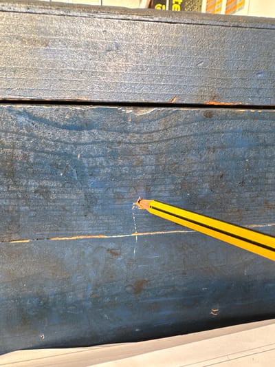A close-up, overhead shot of a weathered, blue wooden surface shows a yellow pencil with a black stripe angled across the lower right portion of the frame. The pencil tip is touching a small, shallow indentation in the wood, suggesting it was used to mark or poke the surface. The wood appears to be constructed from planks, with visible grain lines and signs of wear, including chipped paint and minor discoloration. The background of the image is out of focus, but suggests some printed material or the edge of another object at the top and a white surface at the bottom. There are no people or any other identifiable objects present in the immediate scene. The image does not contain any information related to the city of Mejaniga, Italy.
