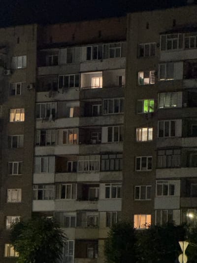 The image shows a nighttime view of a multi-story apartment building, likely in the city of Stryi, Ukraine. The building has a Brutalist architectural style, characterized by its concrete facade and repetitive window patterns. Several windows are illuminated from within, suggesting occupied apartments. One window on the fifth floor from the bottom emits a distinct green light. The lower portion of the building is partially obscured by the dark foliage of trees, and a street light or traffic sign is visible in the bottom right corner. The overall scene depicts a typical urban residential environment after dark.
