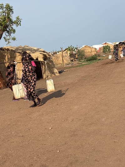 The image shows two women carrying water containers on a dirt road in Shahid Afandi, Sudan. The woman in the foreground is wearing a black and floral patterned dress and is carrying a yoke with two large white water containers. She is walking towards the right side of the image. The second woman, in the background, is partially visible and also appears to be carrying water. They are walking past a small, makeshift structure with a thatched roof, likely a dwelling or shelter. The scene appears to be set during the daytime, with clear skies and bright sunlight casting shadows. The overall setting suggests a rural or semi-urban environment where access to water might be a challenge, requiring people to carry it from a source. There is no visible text in the image.