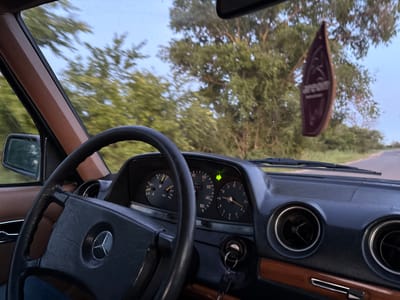The image is a first-person perspective from inside a car, likely a Mercedes-Benz judging by the emblem on the steering wheel. The dashboard is visible, with instruments showing a speed of approximately 90 km/h, a tachometer, and a clock. A green turn signal is illuminated on the dashboard. A dark-colored air freshener, shaped like a canoe or boat, hangs from the rearview mirror. The interior features brown trim on the dashboard and door panel. Outside, trees with green foliage are visible, and the car appears to be on a paved road, suggesting it is in a rural or suburban setting. The sky is visible through the windshield and side windows. The location is identified as Varnița, Moldova.