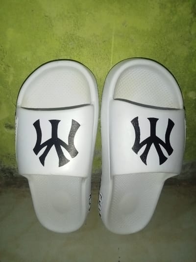 Two white slide sandals are depicted standing upright against a mottled, faded lime-green vertical surface. Both sandals feature a wide forefoot strap and a contoured footbed with a textured, dotted surface. A prominent black stylized emblem, resembling intertwined letters such as "NY" or "W," is centered on the top strap of each sandal. The sandals are aligned parallel to each other, with their toes pointing upwards. Minor, partially obscured text, possibly "PUP," is visible on the lateral side of the right sandal. The background wall exhibits varying shades of green, discoloration, and textural irregularities consistent with an aged or worn surface. No individuals, actions, or specific interactions are present in the scene. The visual evidence in the image is insufficient to identify a specific location within Umsobomvu Local Municipality, South Africa.