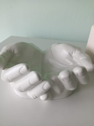 A white, ceramic decorative bowl shaped like cupped hands sits on a white surface against a light green wall. The bowl is hollow in the center, with the fingers and palms of both hands forming its structure. The image is a close-up shot, focusing on the bowl and its immediate surroundings. No people are present in the image. The location is the city of Nizhnetroitskiy, Russia.