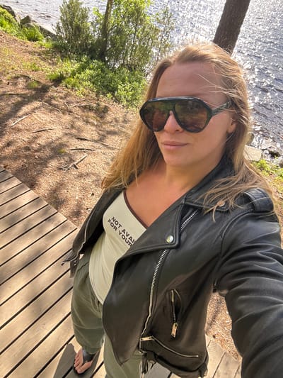 In this high-angle, rotated photograph, a person takes a selfie on a wooden deck near a body of water. The subject, wearing large, dark sunglasses, a black leather jacket, and a light-colored tank top with the text "NOT AVAIL FOR YOUR" visible on it, is positioned in the foreground. Their hair is blonde and windswept, suggesting a breezy environment. The background features shimmering water, lush green foliage, and a tree trunk on the left side of the frame. The wooden deck, made of light-colored planks, dominates the lower right portion of the image. The scene appears to be outdoors during the daytime, indicated by the bright sunlight and strong shadows. The overall mood seems relaxed and casual, perhaps indicating a leisurely activity by the water. The location cues, such as the water, trees, and wooden structures, are consistent with an outdoor recreational area.