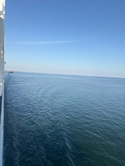 The image depicts a vast expanse of calm, rippling water stretching towards the horizon under a clear, pale blue sky. The water's surface is textured with subtle waves, catching the light and creating a shimmering effect. On the left side of the frame, the sky transitions from a deep blue near the bottom to a lighter shade as it approaches the horizon. A faint, wispy cloud streak is visible in the sky.

In the foreground, a white railing or deck runs horizontally across the bottom of the image. On this structure, a small, orange traffic signal is visible, its light illuminated. Another indistinct dark object, possibly a person, is visible near the traffic signal, and a small dark object is also visible on the far right edge of the railing. The scene suggests an outdoor setting, likely a ferry or a coastal viewing platform, given the railing and the traffic signal.

The lighting indicates it is daytime, possibly late morning or early afternoon, with the sun high in the sky. The overall mood is serene and tranquil, emphasizing the scale of the natural environment. The location is noted as unknown, Lithuania. No visible text is present in the image.